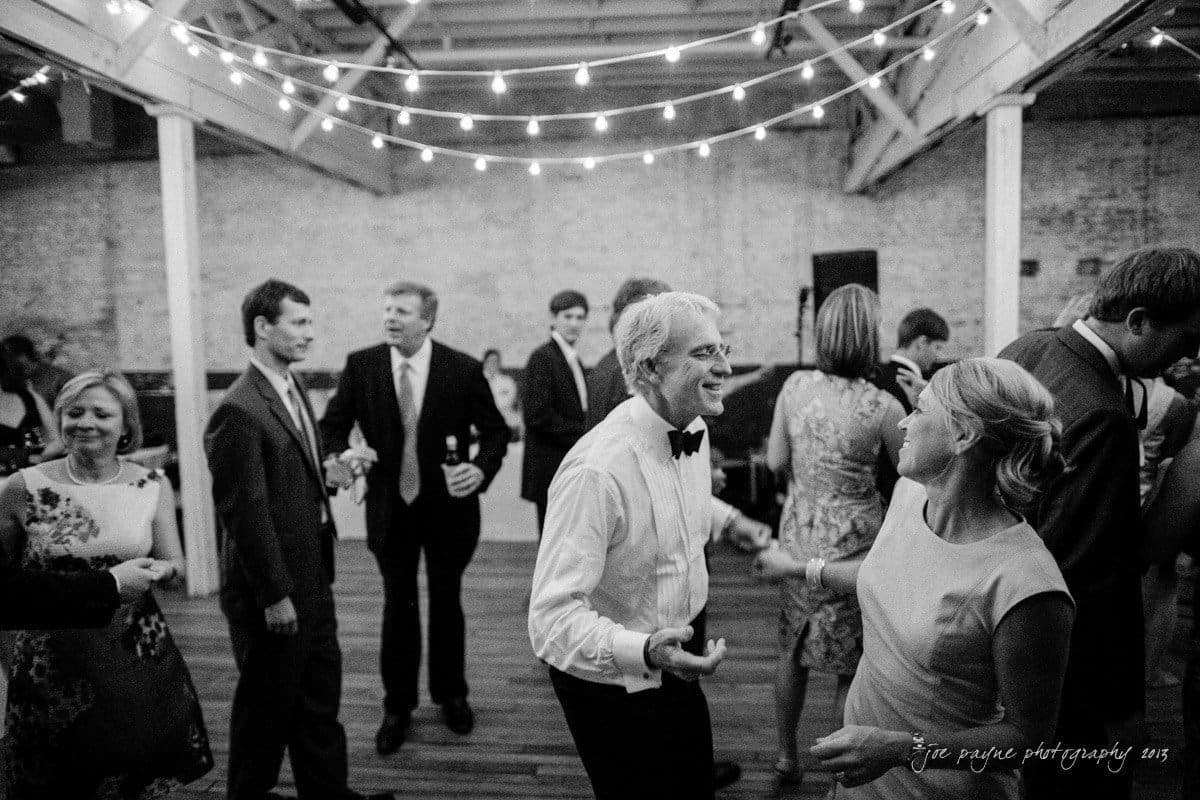 Black and white photo of people dancing at an indoor event. String lights hang overhead, and guests, dressed in formal attire, are enjoying the lively atmosphere.