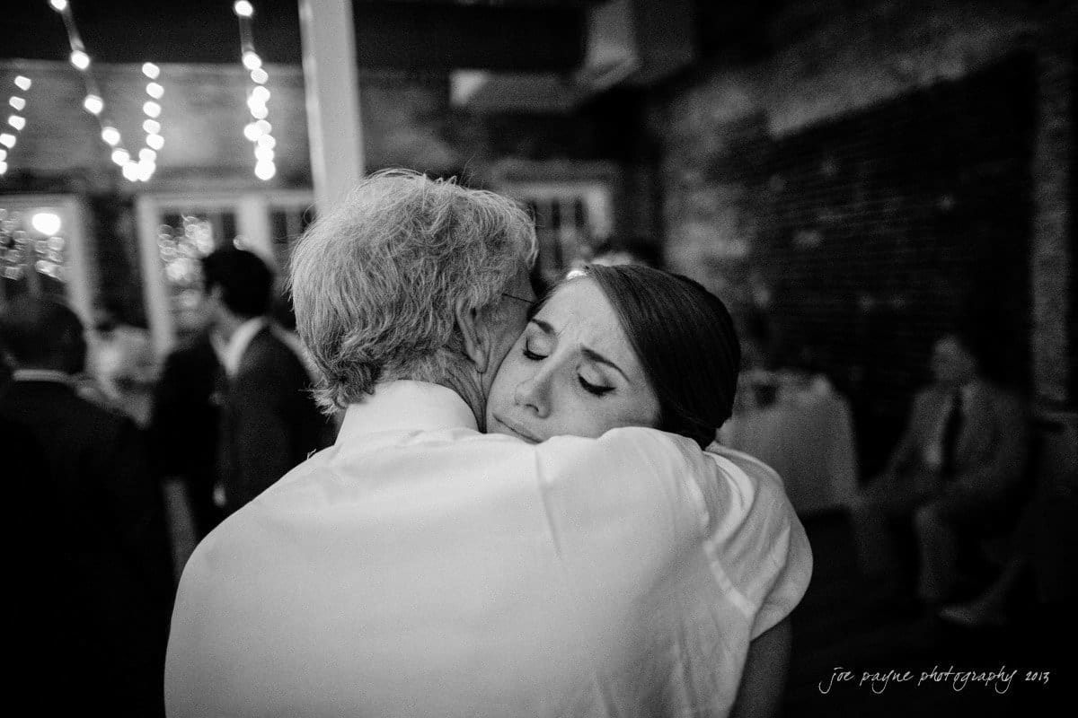 A grayscale image of a man and a woman embracing closely. The woman appears to be emotional, resting her head on the mans shoulder. In the blurred background, there are string lights and people in semi-formal attire.