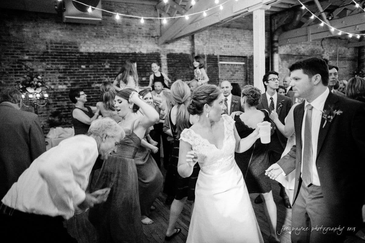 A lively black-and-white photo of a wedding reception. Guests dance energetically under string lights in a rustic venue with brick walls and wooden beams. The bride, holding a drink, and the groom are at the center, enjoying the moment.