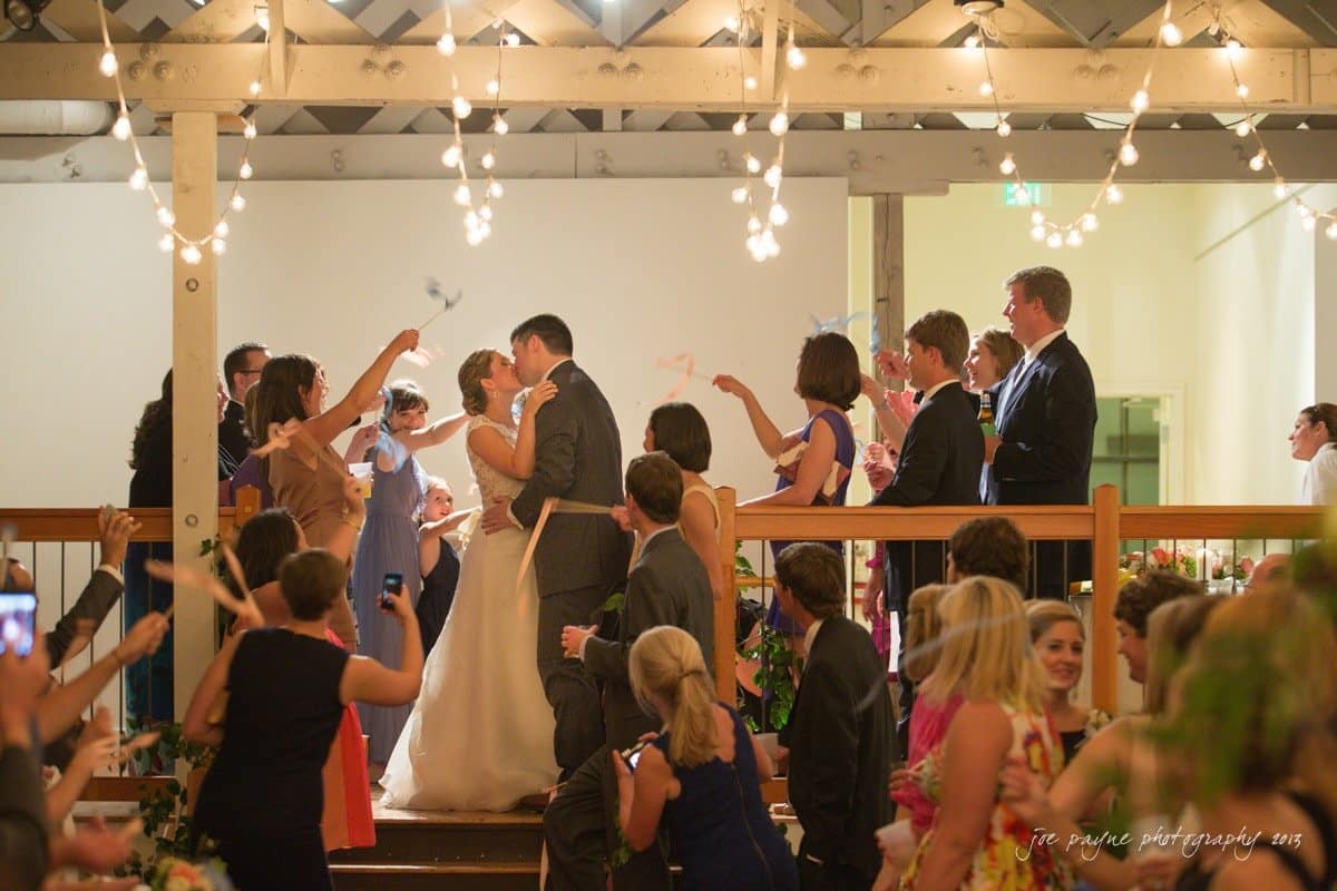 A newlywed couple kisses on a wooden stage surrounded by a cheering crowd. People wave ribbons and celebrate under string lights. The setting appears to be a festive indoor venue.