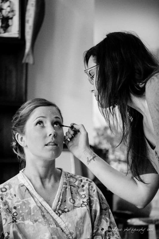 A black-and-white photo shows a woman in a patterned robe sitting while another woman applies mascara to her eyelashes. The makeup artist wears glasses, and the setting appears to be indoors, possibly a home or studio.