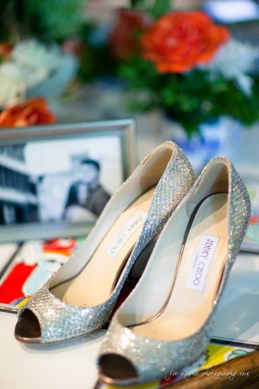 A pair of sparkling Jimmy Choo peep-toe shoes sit on a table. In the background, theres a blurred black-and-white photograph and colorful flowers, including bright orange roses.
