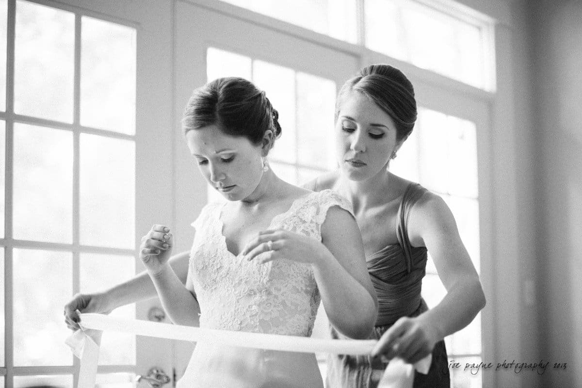 A black and white photo of a bride being helped by a bridesmaid. The bride is wearing a lace wedding dress, and the bridesmaid is tying a sash around her waist. They are standing near a window with soft light filtering in.