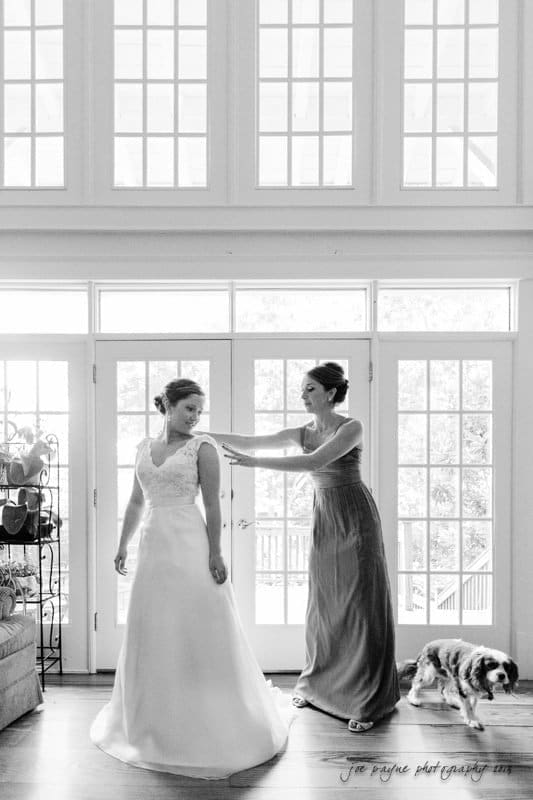 A bride in a white gown stands inside, while another woman in a long dress adjusts her outfit. A dog walks nearby. Sunlight streams through large windows and glass doors in the background.