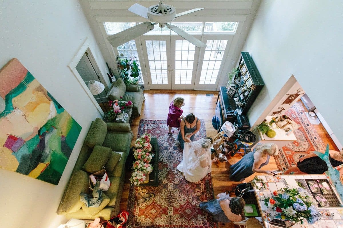 A high-angle view of a living room with a colorful abstract painting on the wall. Several people are gathered around, with a table filled with flowers and decorations. The room features a ceiling fan and large windows with natural light.