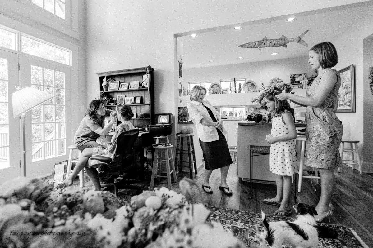 A black-and-white image shows a lively room with several women and a child. One woman styles the childs hair, while another is seated, getting her makeup done. A dog rests on the floor amidst a cozy, decorated space with shelves and wall accents.