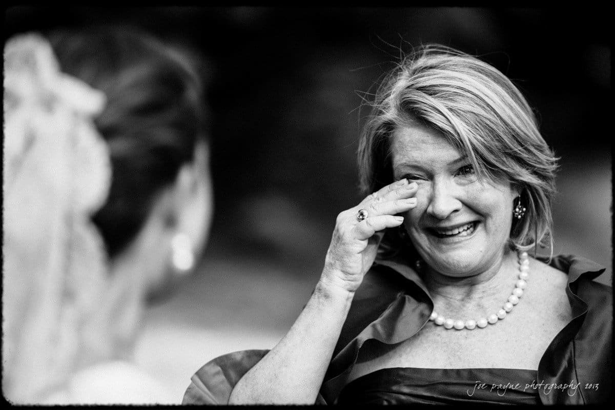 A woman with short hair and a pearl necklace wipes tears from her eyes while smiling, facing another person. The scene is in black and white, conveying an emotional moment.