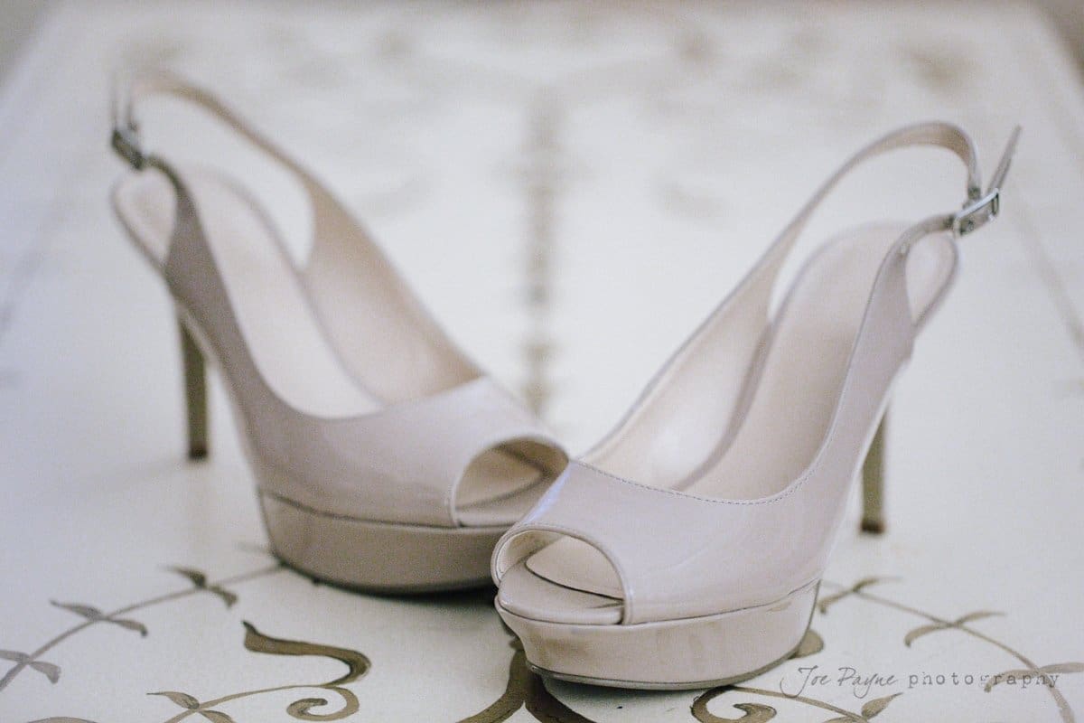 A pair of beige peep-toe slingback high heels sits on an elegant patterned surface. The shoes have a glossy finish and high platform soles.