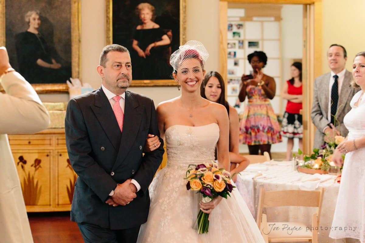 A bride in a white gown and veil walks arm in arm with a man in a suit. She holds a bouquet of flowers. Guests are gathered in the background, and portraits hang on the wall. It appears to be a wedding ceremony.