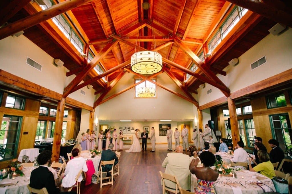 A wedding ceremony inside a wooden hall with a high, vaulted ceiling and large chandeliers. The couple stands in the center, surrounded by a wedding party in formal attire. Guests are seated at round tables on either side.
