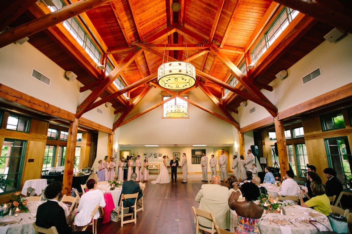 A wedding ceremony inside a wooden hall with a high, vaulted ceiling and large chandeliers. The couple stands in the center, surrounded by a wedding party in formal attire. Guests are seated at round tables on either side.