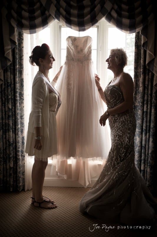 Two women stand by a window admiring a bridal gown suspended between them. One is dressed in a robe and sandals, while the other is in an elegant dress. They are silhouetted by the soft light filtering through the window.