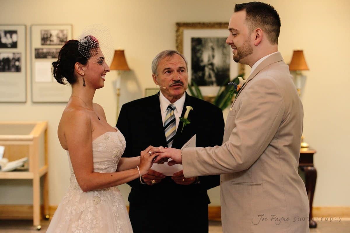 A bride and groom exchange vows in a wedding ceremony. The groom holds the brides hand as they face each other. An officiant stands between them, holding a book. The setting is indoors, with framed pictures on the wall in the background.
