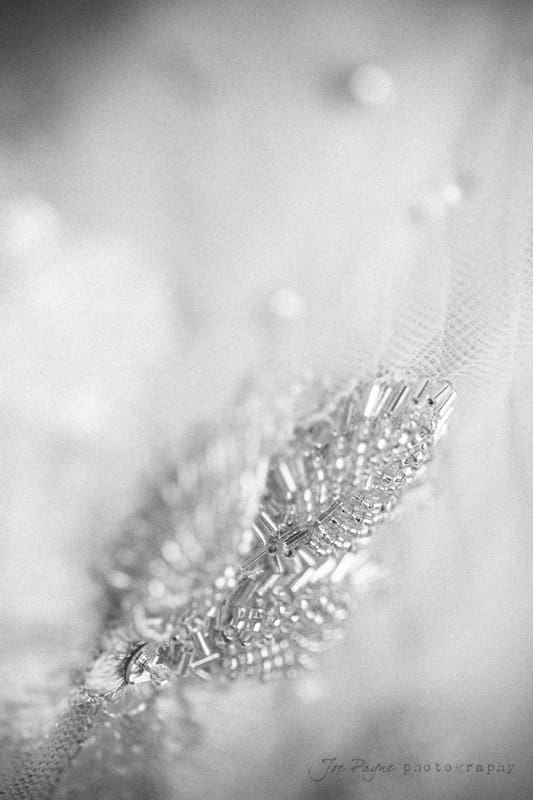 Close-up of intricate beadwork and embroidery on delicate fabric, featuring tiny beads and sequins arranged in an elegant design. The image is in black and white, highlighting the texture and craftsmanship.