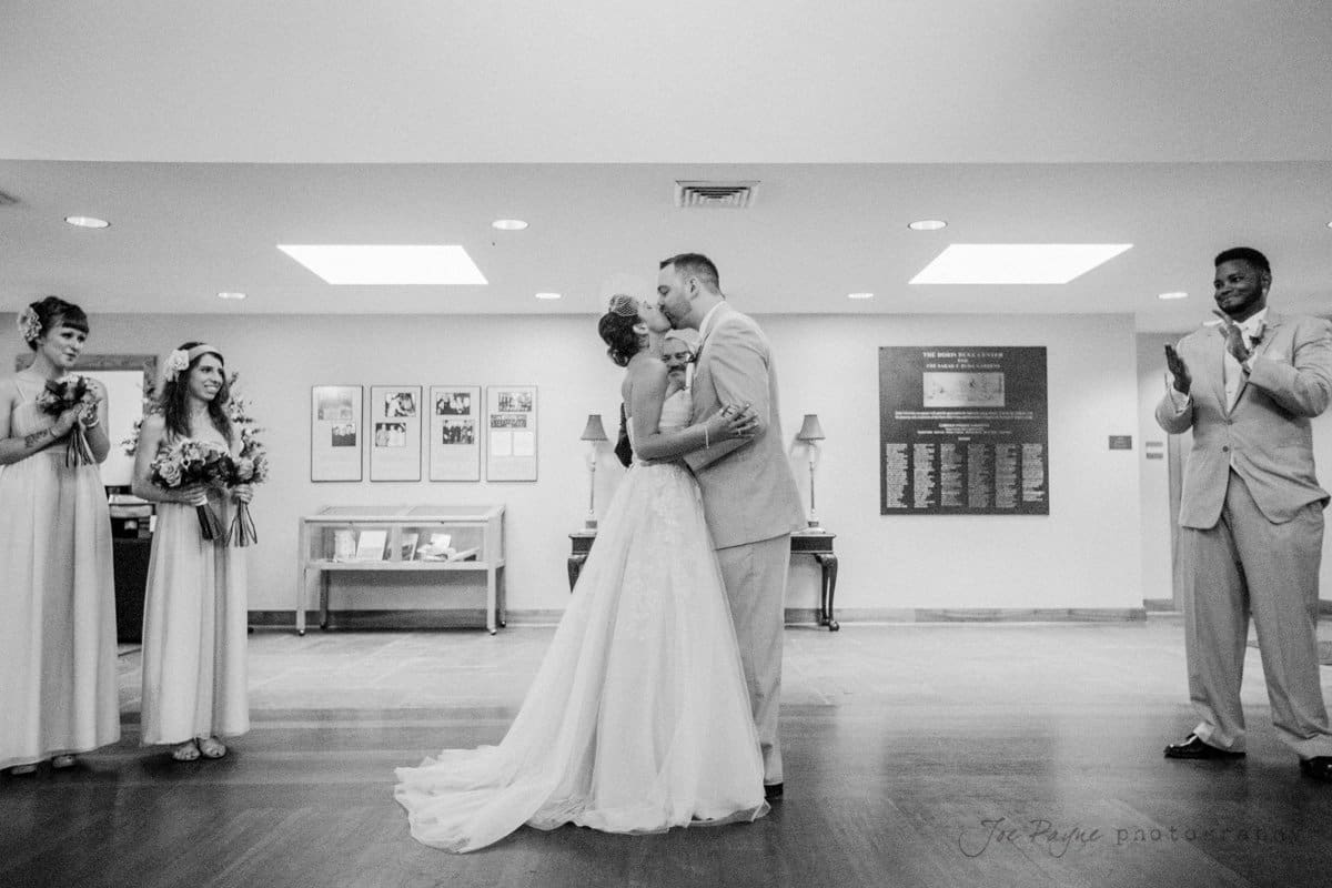 A couple in wedding attire shares a kiss in the center of a room, surrounded by a joyful wedding party. Two bridesmaids on the left and a groomsman on the right clap and smile. The setting is indoors with framed pictures on the walls.