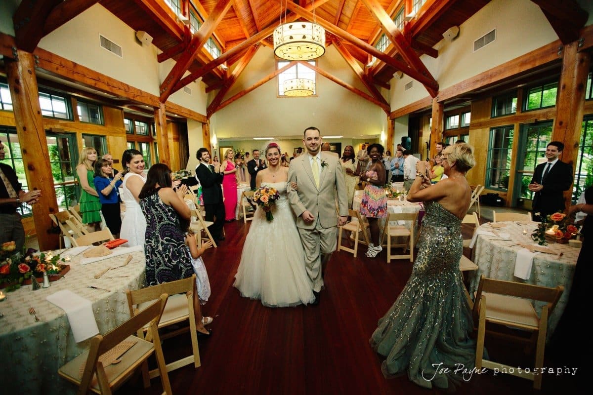 A newlywed couple walks down the aisle in a bright, wooden reception hall. Guests cheer and take photos. The bride holds a bouquet, and the groom smiles. Tables with floral arrangements surround the scene.