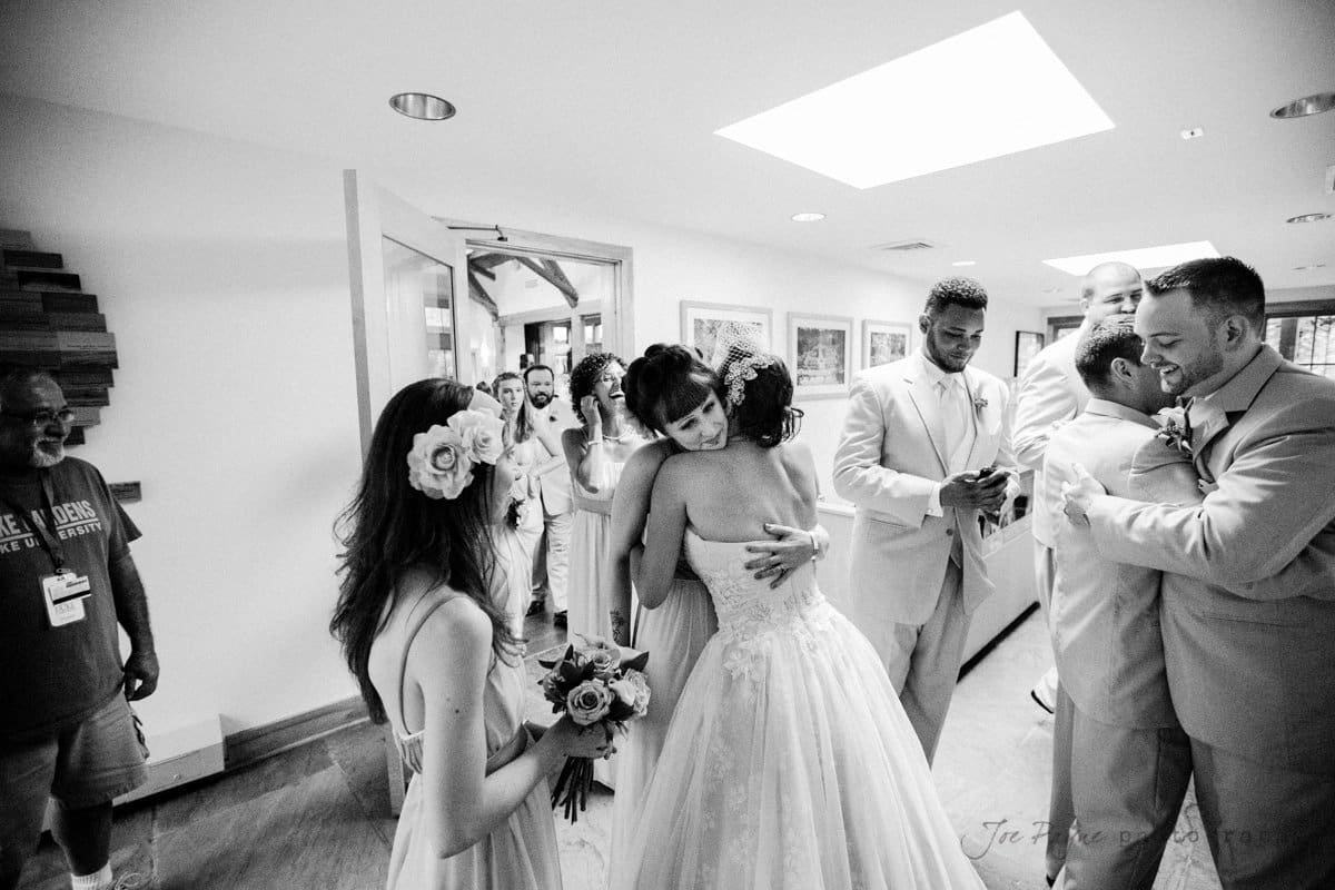 A black and white photo of a wedding reception where the bride and groom are hugging guests. Other attendees, including bridesmaids holding bouquets, are seen mingling in the background. The atmosphere is joyful and celebratory.