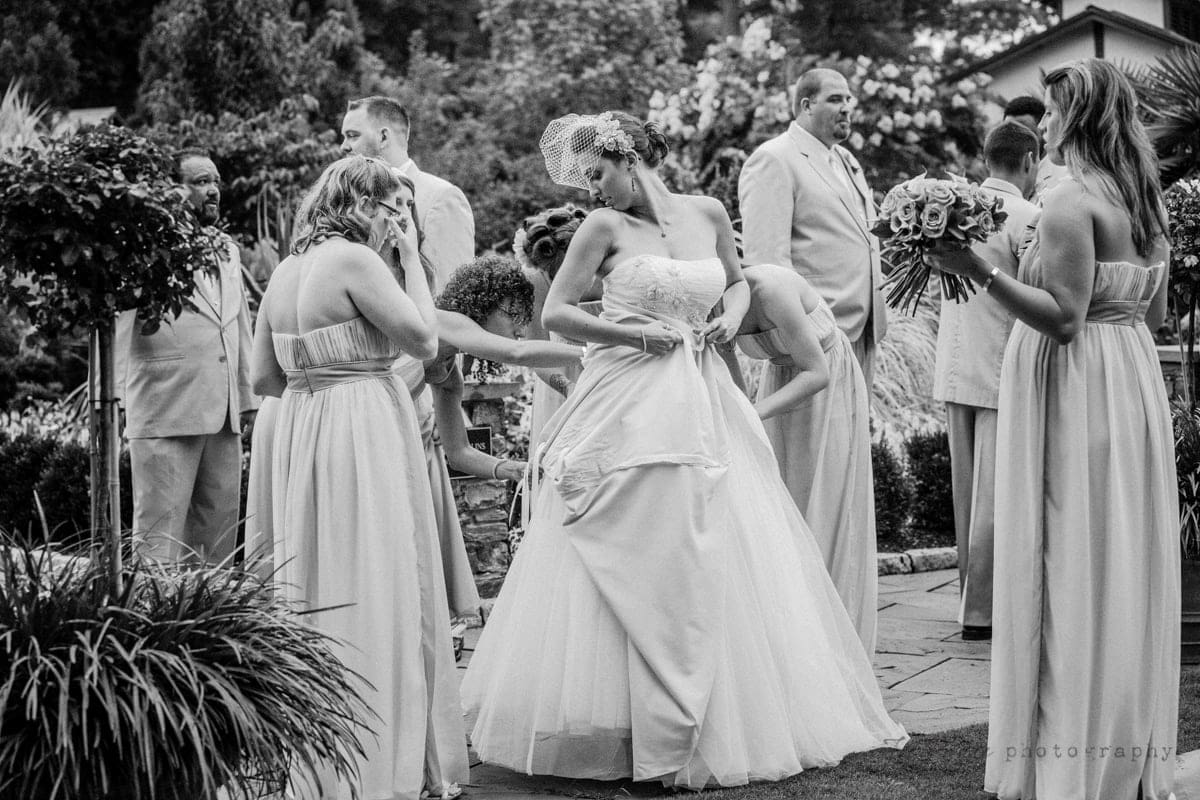 A bride in a flowing gown stands in a garden, surrounded by bridesmaids in pale dresses and groomsmen in suits. One bridesmaid adjusts the brides dress. The scene is lively and the setting is lush with greenery.