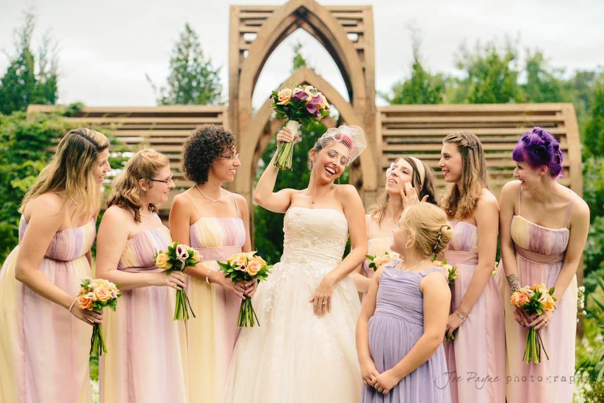 A joyful bride holds a bouquet aloft while surrounded by smiling bridesmaids in pastel dresses. They stand outdoors with greenery and a wooden arch in the background.