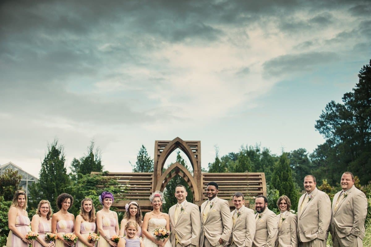 A wedding party stands posed outdoors in front of a wooden arch. The group consists of bridesmaids in light dresses and groomsmen in beige suits, with a child in front. The sky is overcast, and trees surround the scene.