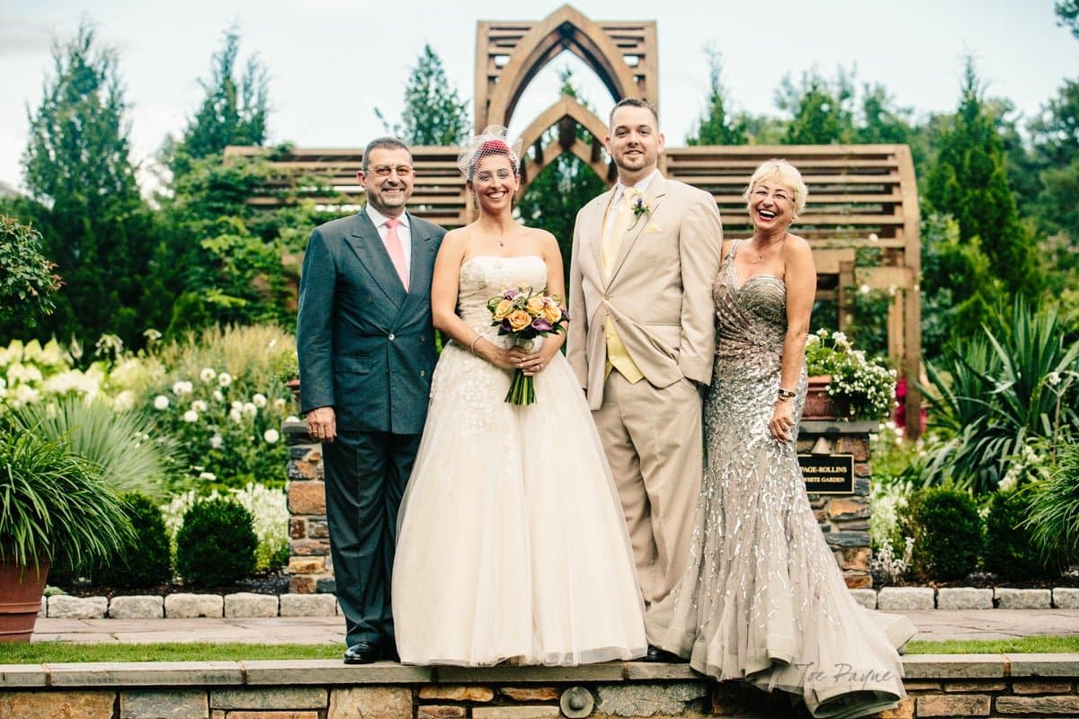 A wedding party posing in a garden. The bride holds a bouquet and wears a white gown, standing next to the groom in a beige suit. Two older adults in formal attire stand on either side, smiling. Lush greenery and a wooden archway are in the background.