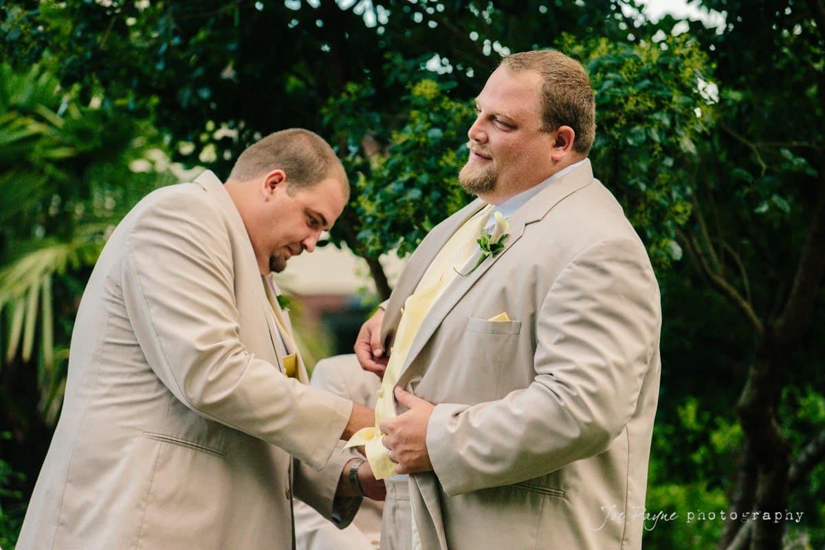 Two men in beige suits adjust their jackets outdoors. One man faces the camera with a thoughtful expression, while the other focuses on adjusting his attire. Green foliage is visible in the background.