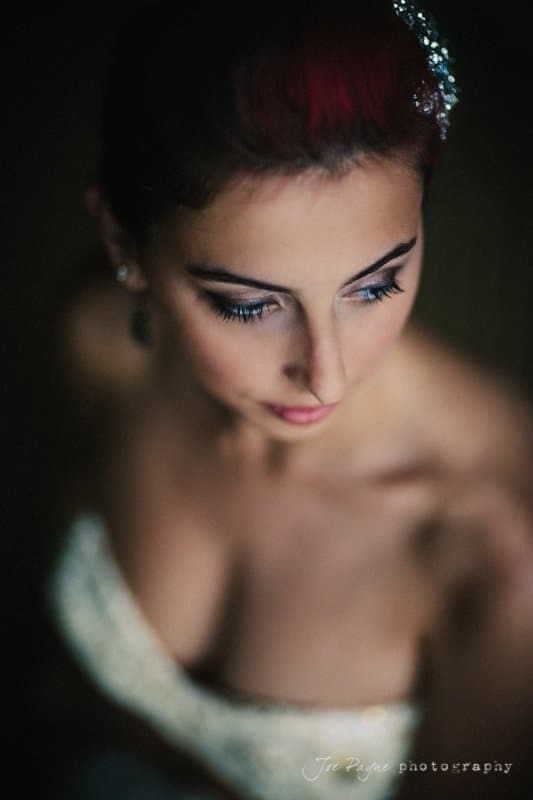 A woman with red-tinted hair and subtle makeup gazes downward. She wears a strapless dress, with light softly illuminating her face and shoulders, against a dark, blurred background.