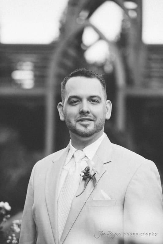 A man in a light-colored suit smiles while standing in an outdoor setting with blurred architectural elements in the background. He wears a tie and boutonniere. The image is in black and white.