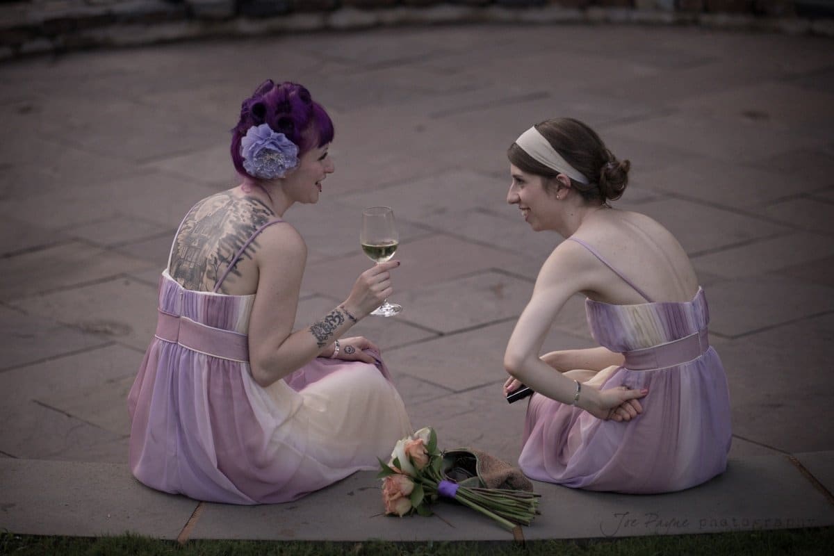 Two women in pastel dresses sit on a stone surface, facing each other. One has purple hair with a large flower accessory and holds a wine glass. A bouquet of roses rests on the ground between them. They appear to be chatting and smiling.