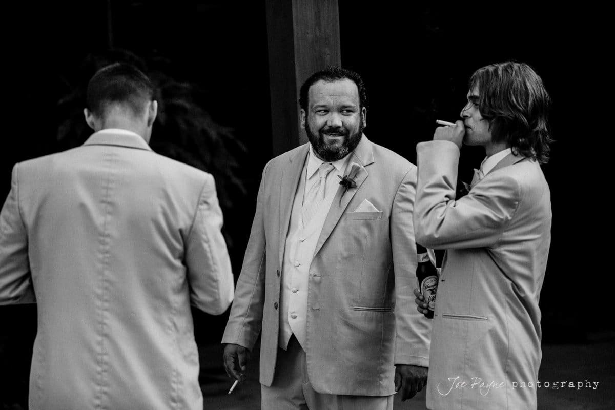 Three men in suits are gathered together. The man in the center is smiling, while the other two men on either side are smoking. The setting appears to be formal, possibly at an outdoor event. The photo is in black and white.