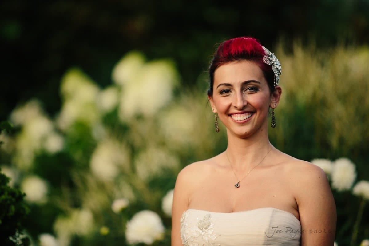 A woman in a strapless white dress with a floral accessory in her hair smiles while standing in front of a lush, blurred green and white floral background.