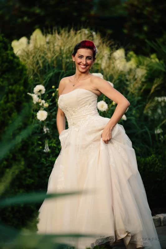 A bride in a strapless white wedding gown stands outdoors in a lush garden. She has a radiant smile and holds her dress with one hand. The background features greenery and white flowers.
