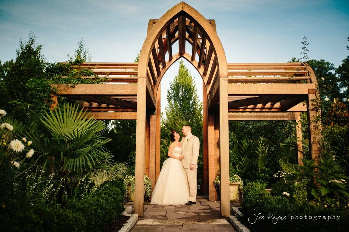 A couple in wedding attire stands under a wooden archway in a lush garden. The bride wears a white dress and the groom a light suit. The scene is filled with greenery, flowers, and clear skies, creating a romantic and serene atmosphere.