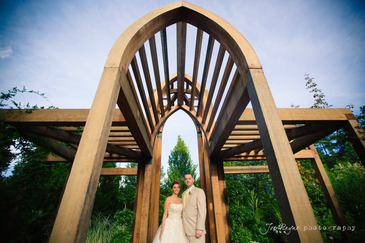 A couple stands under a wooden pergola with arched beams, surrounded by lush greenery. The bride wears a strapless white dress, and the groom is in a light-colored suit. The sky is clear and blue, adding to the serene outdoor setting.