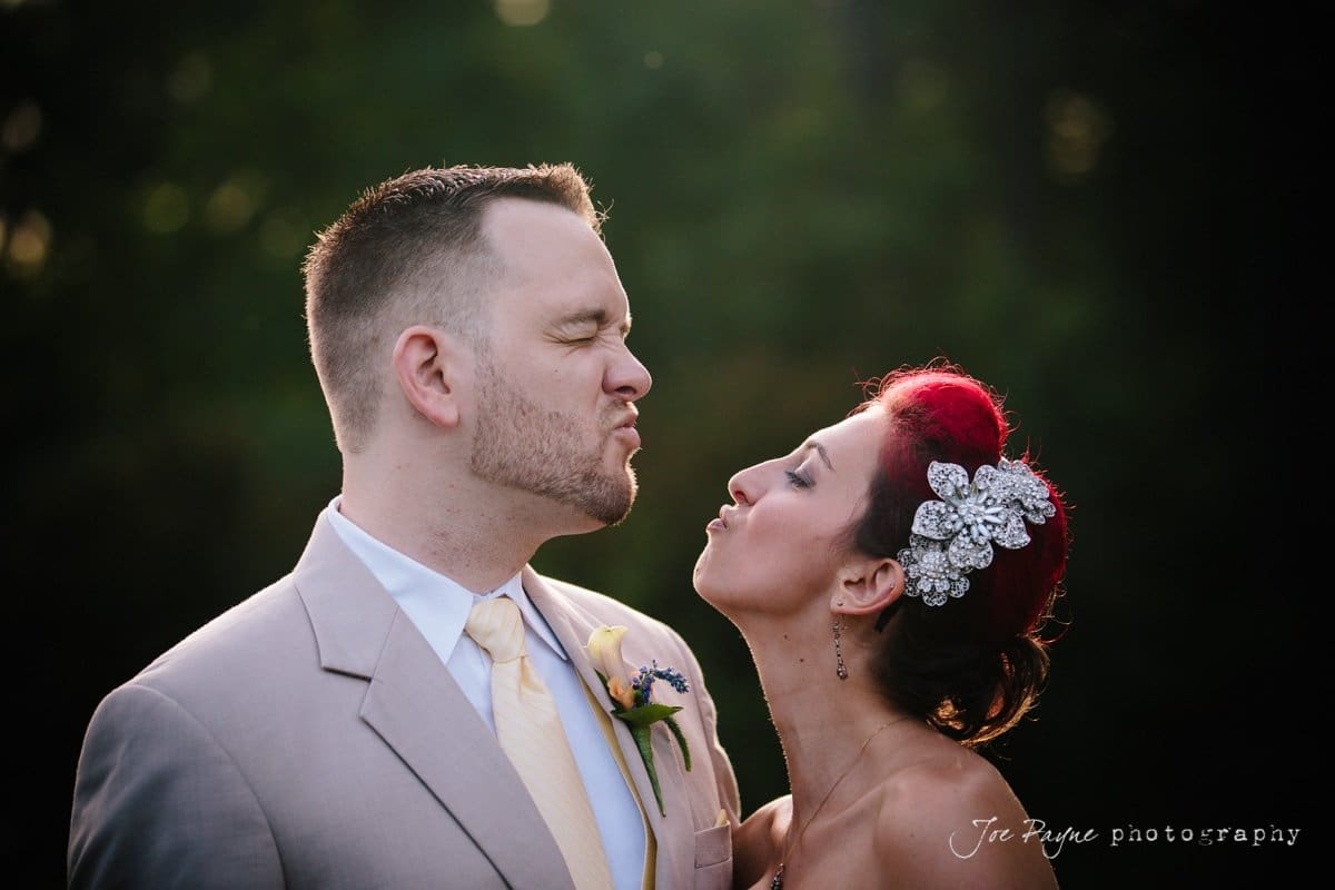 A groom in a beige suit playfully purses his lips as a bride with red hair and decorative hairpieces leans towards him as if to kiss. They stand outdoors, surrounded by dark greenery. The photo is by Jon Payne Photography.