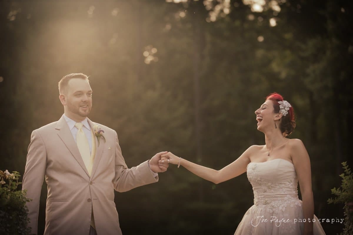 A bride and groom stand outdoors, holding hands and smiling joyfully. The bride wears a strapless white gown and a hair accessory, while the groom is in a beige suit with a tie. Sunlight filters through the trees in the background.