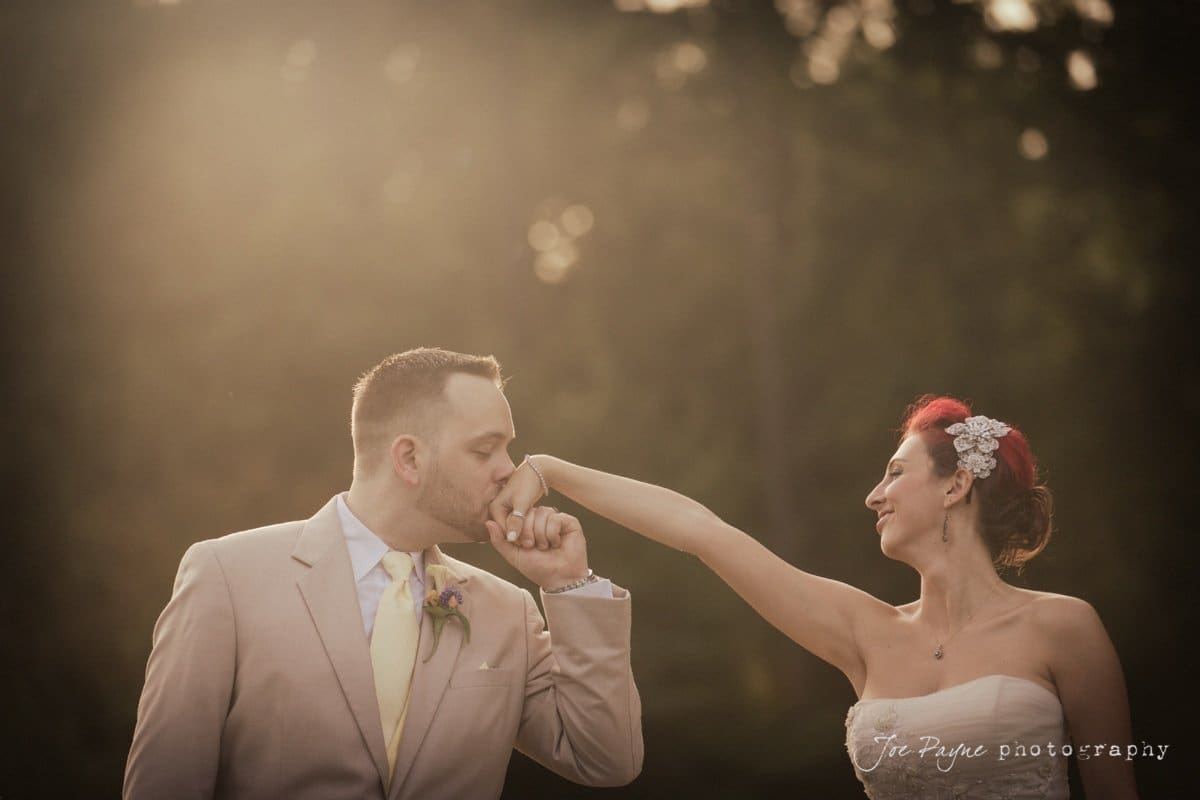 A groom in a beige suit kisses the hand of a bride in a strapless white gown and floral hairpiece. They stand outdoors with a blurred dark green background and soft sunlight filtering through.