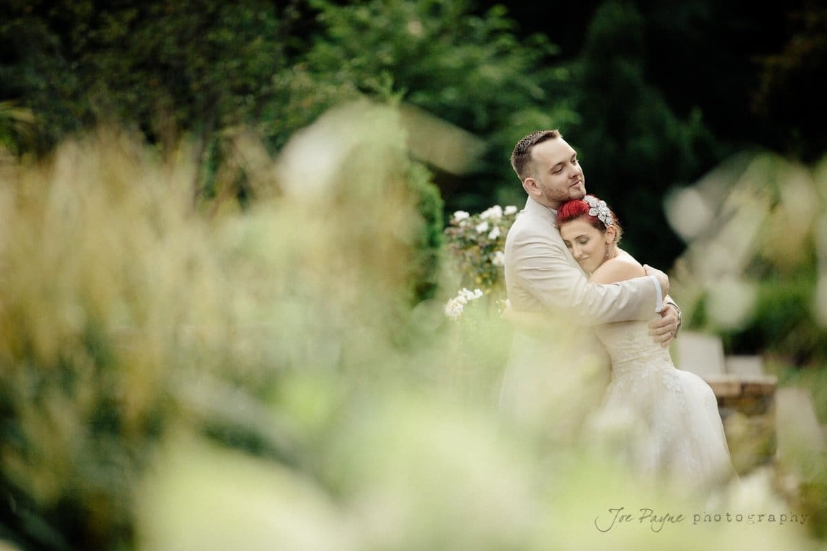 A bride and groom embrace tenderly in a garden setting. The bride wears a floral headpiece, and the groom is in a light suit. The foreground is softly blurred with greenery, creating a dreamy and romantic atmosphere.