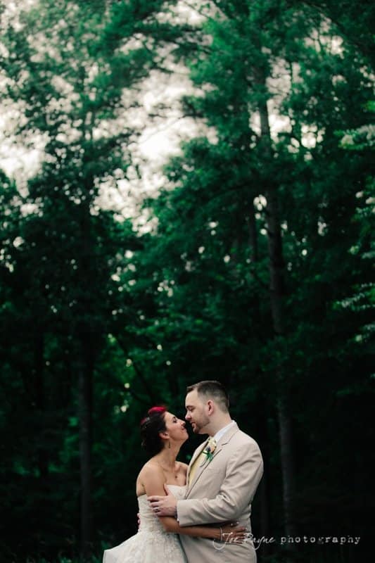 A bride and groom embrace under tall, lush trees. The bride wears a white dress, and the groom is in a light suit. The setting is serene and green, conveying a romantic atmosphere.