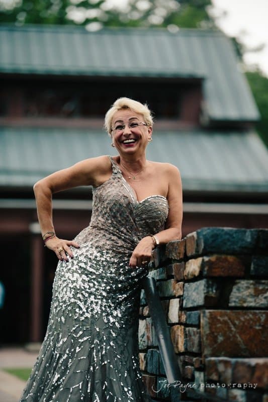 A smiling person in a sparkling, one-shouldered gown leans casually against a stone wall. The background shows a building with a gray metal roof.