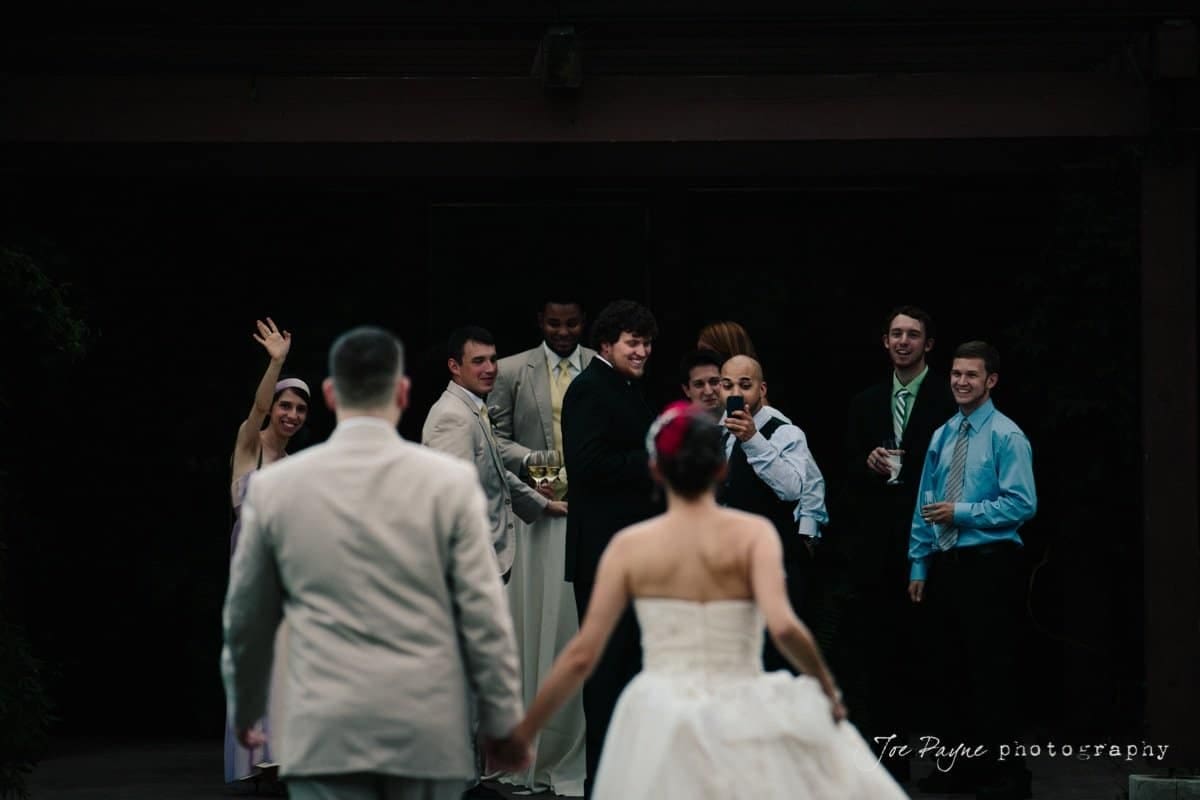 A bride and groom, seen from behind, walk toward a group of smiling groomsmen dressed in suits and ties. One woman playfully waves in the background. The atmosphere is joyful and celebratory.
