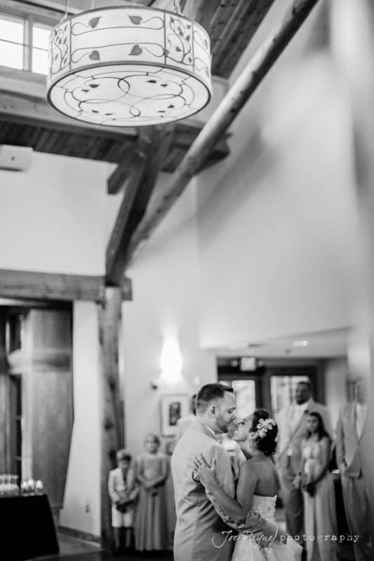 A bride and groom share their first dance in a warmly lit room with wooden beams. Guests watch the couple in the background. A decorative ceiling light is visible above.