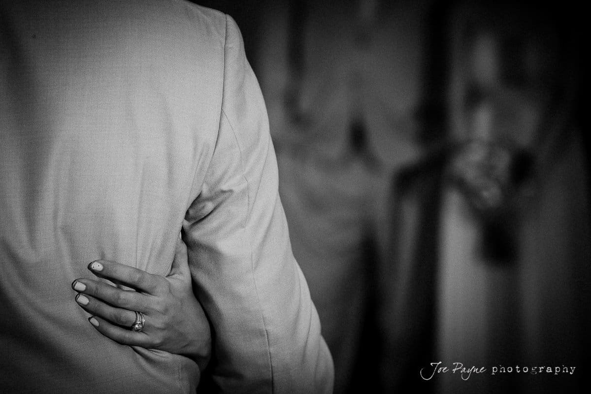 Black and white photo showing a close-up of a couple embracing. A woman’s hand with a ring rests on the back of a person in a suit.