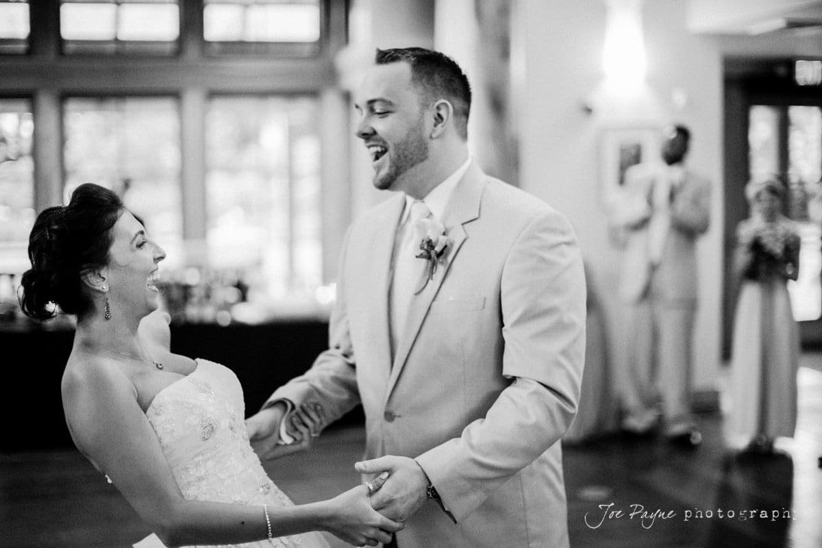 A bride and groom joyfully dance together at their wedding reception. They are smiling and holding hands, surrounded by a softly lit venue. Guests in the background watch the couple. The setting is elegant and festive.