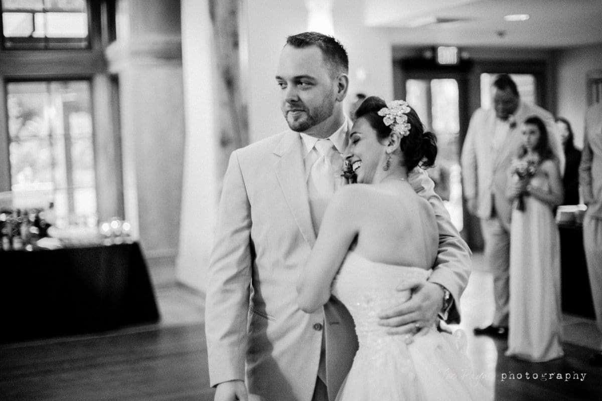 A bride and groom share an embrace in a warmly lit room, surrounded by guests. The bride wears a strapless dress and a floral hairpiece, while the groom dons a light suit. The atmosphere is joyful and intimate.