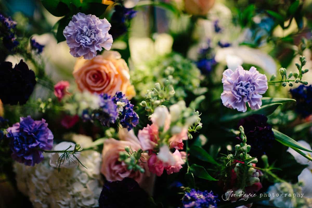 A colorful bouquet featuring purple carnations, peach roses, and various green foliage, beautifully arranged. The flowers are vibrant and fresh, captured in a well-lit setting, creating an artistic and lively floral display.