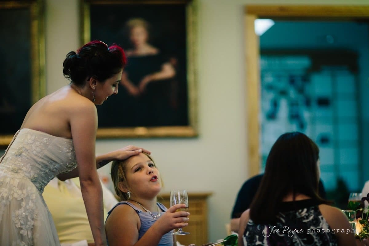 A woman in a strapless dress gently touches the head of a seated girl holding a glass. They are indoors, with framed art hanging on the wall behind them. Other people are seated nearby, partially visible in the scene.