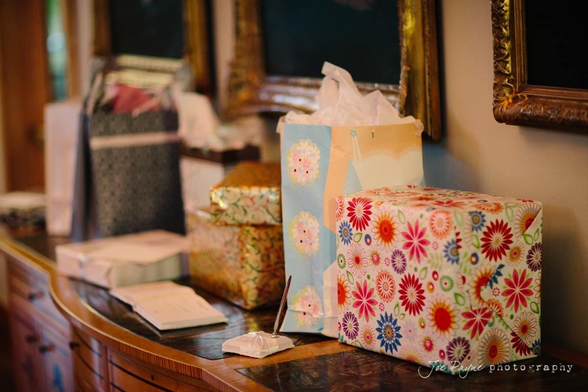 Gift bags and wrapped presents are lined up on a wooden table. The bags and boxes are colorful with floral and decorative patterns. Framed artworks are visible in the background on the wall.