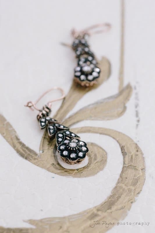 Ornate black and silver earrings with floral patterns rest on a decorative, vintage-style gold and white surface. The intricate design features small gemstones, enhancing their elegant appearance.