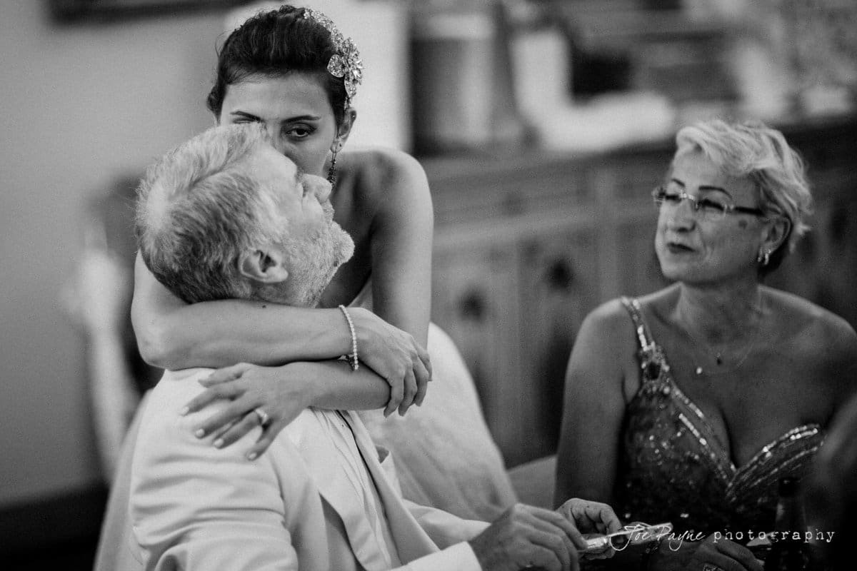 A black and white photo shows a bride hugging an older man, possibly her father, while an older woman, in a sequined dress, sits nearby. The scene appears tender and emotional.